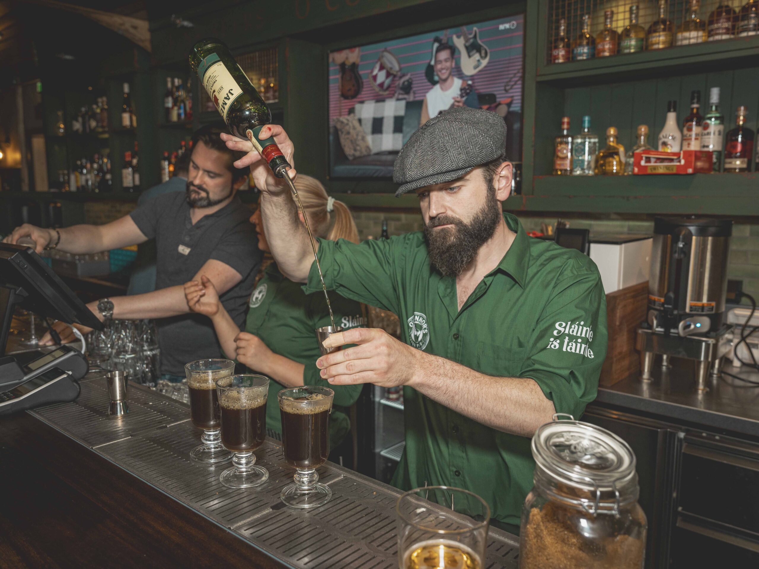 To menn og en kvinne som jobber som bartendere, mannen i forgrunnen lager en drink hvor han heller fra en flaske ned i et glass.